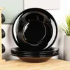 A stack of black plates on a wooden surface, with a plant in a pot in the background. Podgląd zdjęcia produktu