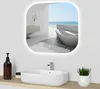 A modern bathroom vanity with a sink, faucet, and a large illuminated mirror reflecting a luxurious bathroom with a bathtub and ocean view.