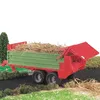A toy farm trailer filled with straw, shown in a diorama setting with miniature trees and bushes. Náhled obrázku produktu