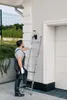 A man is using a ladder to reach a security camera mounted on the side of a building. Voir les images du produit