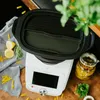 A food processor with a steamer basket attachment, surrounded by pasta, lemons, and herbs. Náhľad obrázku produktu