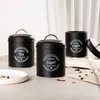 Three black metal canisters for tea, sugar, and coffee are arranged on a table with some dried tea leaves and petals scattered around. Produktbild-Vorschau