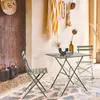 An outdoor patio scene with a bistro table and chairs set up on a stone floor, decorated with hanging items and lanterns. Produktbild-Vorschau