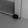 A close-up view of the bottom corner of a glass shower door with black hardware, resting on a gray floor against a gray tiled wall. Produktbild-Vorschau