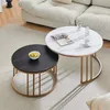 Two round coffee tables, one white marble-effect and one black marble-effect, are placed on a rug in front of a sofa. The white table has a magazine, a cup and saucer, and two bottles of water on it. The black table has a bowl of fruit on it. The tables ha Produktbild-Vorschau
