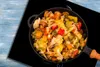 A close-up overhead view of a pan filled with stir-fried vegetables on a black induction hob, with a blue wooden surface visible to the left. Produktbild-Vorschau