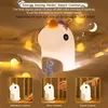 A cute unicorn-shaped night light with a digital clock display, shown in a cozy, dimly lit bedroom setting with fairy lights. Three smaller inset images demonstrate different control methods: vibration, voice, and touch. Produktbild-Vorschau