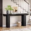 A black wooden console table is placed in a hallway next to a staircase. The table has a vase with green leaves, a framed picture, and two candlesticks on top. A woven basket with a pillow is placed underneath the table. Produktbild-Vorschau