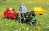 A young boy sits on a green toy tractor with a red trailer filled with flowers, in a field of dandelions. Produktbild-Vorschau