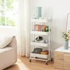 A white rolling cart with four shelves filled with various items, placed next to a sofa and a cabinet with flowers. Voir les images du produit