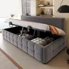A dark gray upholstered bed frame with the mattress lifted to reveal storage space underneath. The bed is in a room with blinds and shelves in the background. Anteprima dell’immagine prodotto