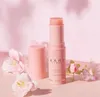 A pink Kahi Wrinkle Bounce Multi Balm stick is displayed upright, with its cap resting beside it. Delicate pink flowers are arranged around the product, creating a soft and aesthetic presentation. Náhled obrázku produktu