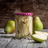 A jar of preserved pears with whole pears and pear slices on a wooden surface. Voir les images du produit