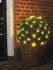 A spherical bush decorated with warm white LED lights, placed in a wicker basket next to a brick wall and a dark door. Produktbild-Vorschau