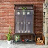 A mini greenhouse on a stand next to a brick wall, with gardening supplies and firewood nearby. Voir les images du produit