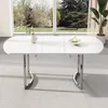 A white dining table with silver legs is shown in a well-lit room with a large window overlooking a garden. Náhled obrázku produktu