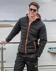 A man wearing a black puffer jacket with orange accents stands by a railing overlooking a body of water with a boat in the background. Produktbild-Vorschau