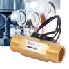 A brass flow meter with wires attached, set against a blurred background of industrial gauges and pipes. Produktbild-Vorschau