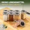 Several clear plastic spice containers with gray lids are arranged on a wooden surface. One container is tipped over, spilling pink salt onto the surface. A green leaf logo with text is in the bottom left corner. Produktbild-Vorschau