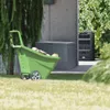A green wheelbarrow filled with wood is positioned in front of a grey storage box and a black compost bin, set in a garden environment. Náhled obrázku produktu