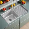 A stainless steel kitchen sink with a faucet and a draining board, surrounded by fruits and vegetables on a countertop. Anteprima dell’immagine prodotto