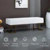 A white upholstered bench with gold metal legs is placed in a modern living room setting. The background features a framed artwork and a tall white vase. Náhľad obrázku produktu