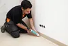 A man is kneeling and applying sealant to the base of a wall. Produktbild-Vorschau