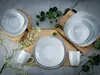 A set of white porcelain dishes arranged on a textured surface with decorative greenery and flowers. Produktbild-Vorschau