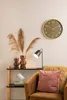 A clock on a wall above a shelf with decorative items and a chair with pillows. Produktbild-Vorschau
