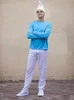 A person wearing a blue shirt and white pants with a white pointed hat, standing in front of a textured wall. Produktbild-Vorschau
