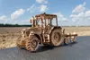 A wooden model tractor with a plow attachment sits on a dark surface in front of a blurred field and sky. Náhľad obrázku produktu