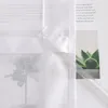 A close-up view of a white sheer curtain with a printed floral design, partially covering a magazine with text and a succulent plant. Produktbild-Vorschau