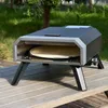 A compact gas pizza oven with a pizza stone inside, placed on a wooden table outdoors with greenery in the background. Produktbild-Vorschau