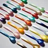 A collection of colorful acrylic spoons arranged diagonally on a light gray background. Produktbild-Vorschau
