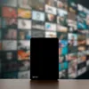 A black electronic device with the brand name 'Vayox' on the bottom left corner, placed on a wooden surface with a blurred background of multiple screens. Podgląd zdjęcia produktu