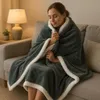 A woman is sitting on a couch, wrapped in a cozy blanket, with a lamp on a side table. Produktbild-Vorschau