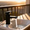 A black faucet is running water into a white sink on a deck with a railing and a sunset in the background. Produktbild-Vorschau