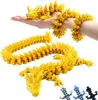A person's hand and arm are holding a yellow, segmented dragon figurine. Another segmented dragon figurine is laid out on a white surface below. Smaller, multi-colored dragon figurines are in the bottom right corner. Produktbild-Vorschau