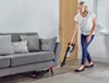 A woman is vacuuming the floor next to a sofa. Produktbild-Vorschau