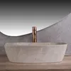 A beige marble vessel sink with a copper faucet is shown in a bathroom setting with a textured gray wall and a wooden countertop. Náhled obrázku produktu