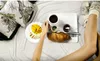 A person's legs are visible as they hold a cup of coffee over a tray with breakfast items on a bed. Produktbild-Vorschau
