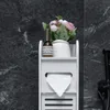 A white bathroom storage unit with a toilet paper dispenser and a shelf holding a decorative plant and a small bottle, set against a dark, textured wall. Náhled obrázku produktu