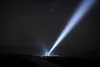 A person stands in a dark, open landscape, illuminated by a powerful flashlight beam pointing upwards into a starry night sky. Podgląd zdjęcia produktu