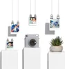 A Fujifilm instax SQUARE SQ 1 camera is displayed on a white pedestal, surrounded by hanging photos and a potted plant. Anteprima dell’immagine prodotto