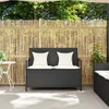 A black poly rattan garden bench with white cushions sits on a patio in front of a bamboo fence. Anteprima dell’immagine prodotto