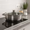 A stainless steel pot with a glass lid sits on a modern induction stovetop, with jars of ingredients and a potted herb in the background. Produktbild-Vorschau