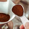 A hand holding a measuring spoon filled with coffee grounds over a coffee filter. Produktbild-Vorschau