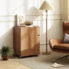 A wooden cabinet with a door and wheels, next to a brown leather chair and a floor lamp, with a plant in the foreground. Produktbild-Vorschau