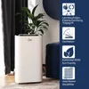 A white mobile air conditioner unit is shown in a room with a plant on top, next to a blue ottoman and dark curtains. Produktbild-Vorschau