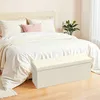 A cream-colored storage ottoman is placed in front of a bed with white bedding and a light pink pillow. Produktbild-Vorschau